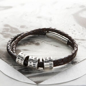 May include: A brown leather bracelet with four silver beads. The beads are engraved with the words "YOU DARE", "XXX", "I LOVE YOU", and "11.24.15".