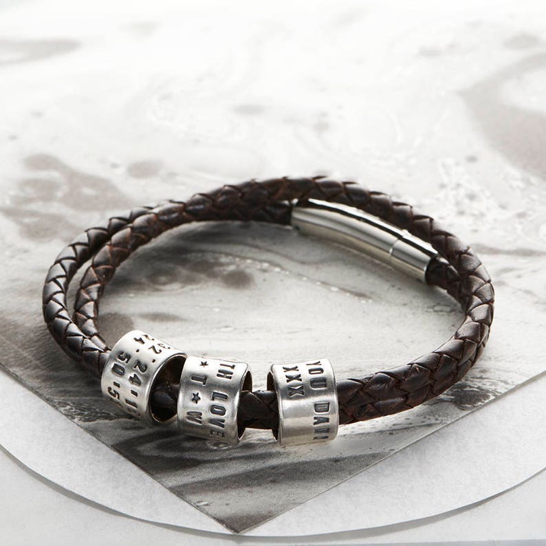 May include: A brown leather bracelet with four silver beads. The beads are engraved with the words "YOU DARE", "XXX", "I LOVE YOU", and "11.24.15".