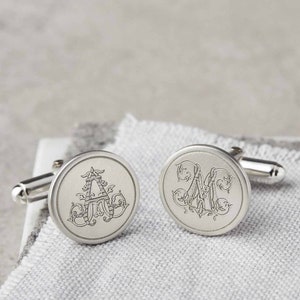Può includere: Un paio di gemelli d'argento con iniziali incise. I gemelli sono rotondi e hanno un design decorativo attorno alle iniziali.