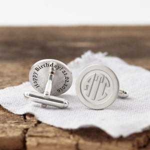 May include: Silver cufflinks with a circular design. One cufflink is engraved with the text "Happy Birthday! 21.02.2016" and the other cufflink is engraved with the monogram "ATC".