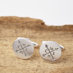 May include: A pair of silver cufflinks with a compass design. Each cufflink features a circle with four arrows pointing to the cardinal directions, with the letters "C", "S", and "02" or "15" and "20" inscribed within the circle.