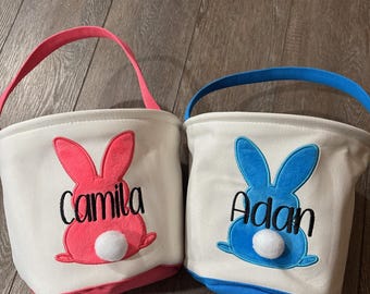 May include: Two personalized Easter baskets with bunny ears and fluffy tails. One basket is pink with the name "Camila" and the other is blue with the name "Adam".