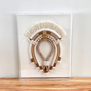 May include: A framed wall hanging with a woven macrame design in beige, brown, and white. The macrame is in the shape of a rainbow with a fringe at the top.