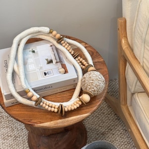 May include: A white rope with wooden beads and a woven ball sits on a wooden stool with a book titled "Minimalista" by Joanna Gaines. The stool is on a beige rug next to a wooden chair with a white cushion.