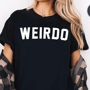 May include: Black t-shirt with the word "WEIRDO" in bold, white capital letters. The shirt is worn with a plaid flannel shirt. A small circle in the upper right corner indicates color options.