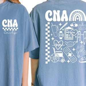 K&ouml;nnte beinhalten: Hellblaues T-Shirt mit den wei&szlig;en Buchstaben "CNA" &uuml;ber einem Schachbrettmuster. Auf der R&uuml;ckseite des Shirts befindet sich eine wei&szlig;e Grafik mit einem Regenbogen, medizinischen Symbolen und den Buchstaben "CNA".