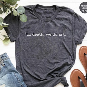May include: Dark grey t-shirt with the text "'til death, we do art." The shirt has a crew neck and rolled-up sleeves. A pair of brown sandals and a pair of blue jeans are visible in the image.