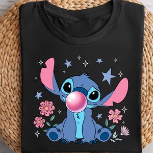 May include: Black t-shirt featuring a cartoon character with blue fur, large ears, and a pink bubble gum bubble. The design includes pink flowers, stars, and the character's name. The shirt is displayed on a woven, circular mat.