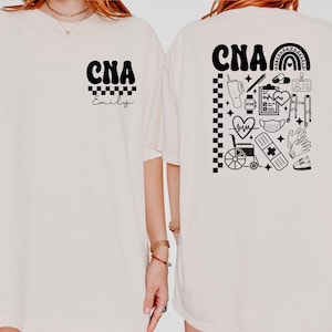 May include: Cream-colored t-shirt with black text and graphics. The front features "CNA" above a checkered pattern and the name "Emily." The back displays "CNA" with a rainbow and medical-related icons. A comfortable, casual style.