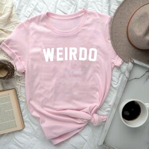 May include: A light pink t-shirt with the word "WEIRDO" printed in white block letters. The shirt is knotted at the bottom. A gray hat, a tray with a cup of coffee, and a book are also in the image.