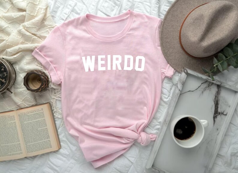 May include: A light pink t-shirt with the word "WEIRDO" printed in white block letters. The shirt is knotted at the bottom. A gray hat, a tray with a cup of coffee, and a book are also in the image.