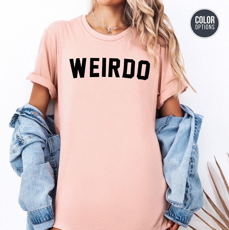 May include: A light pink t-shirt with the word "WEIRDO" printed in bold black letters. The shirt has rolled-up sleeves and is paired with a light blue denim jacket.