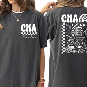 K&ouml;nnte beinhalten: Dunkelgraues T-Shirt mit wei&szlig;em Text und Grafiken. Auf der Vorderseite steht "CNA" &uuml;ber einem Schachbrettmuster und dem Namen "Emily". Auf der R&uuml;ckseite steht "CNA" mit einem Regenbogen und medizinischen Symbolen, darunter ein Herzmonitor, ein Rollstuhl und ein Verband.