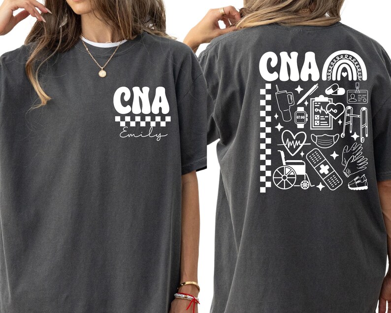 K&ouml;nnte beinhalten: Dunkelgraues T-Shirt mit wei&szlig;em Text und Grafiken. Auf der Vorderseite steht "CNA" &uuml;ber einem Schachbrettmuster und dem Namen "Emily". Auf der R&uuml;ckseite steht "CNA" mit einem Regenbogen und medizinischen Symbolen, darunter ein Herzmonitor, ein Rollstuhl und ein Verband.