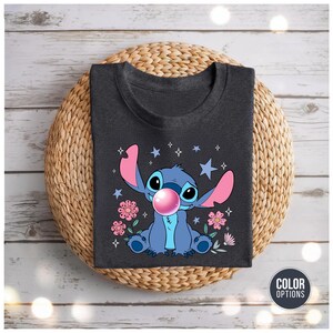 May include: Dark gray t-shirt featuring a cartoon character blowing a bubblegum bubble. The character is blue with large ears and is surrounded by pink flowers and stars. The shirt is displayed on a woven mat.