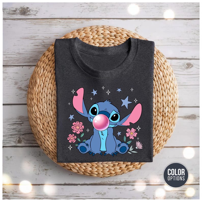 May include: Dark gray t-shirt featuring a cartoon character blowing a bubblegum bubble. The character is blue with large ears and is surrounded by pink flowers and stars. The shirt is displayed on a woven mat.