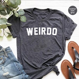 May include: Dark grey heather t-shirt with the word "WEIRDO" printed in bold white letters. The shirt has a crew neck and short sleeves. The image also shows a pair of brown sandals and a pair of blue jeans.