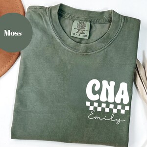 K&ouml;nnte beinhalten: Ein moosgr&uuml;nes T-Shirt mit dem wei&szlig;en Schriftzug "CNA" in Retro-Schrift, &uuml;ber einem schwarz-wei&szlig;en Schachbrettmuster. Der Name "Emily" steht darunter in Schreibschrift. Das Shirt ist aus weichem Material.