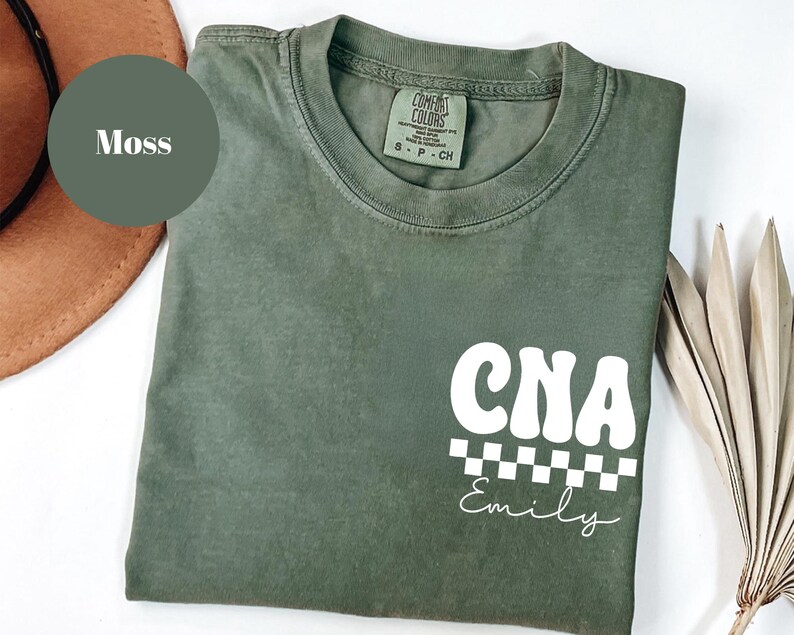 K&ouml;nnte beinhalten: Ein moosgr&uuml;nes T-Shirt mit dem wei&szlig;en Schriftzug "CNA" in Retro-Schrift, &uuml;ber einem schwarz-wei&szlig;en Schachbrettmuster. Der Name "Emily" steht darunter in Schreibschrift. Das Shirt ist aus weichem Material.