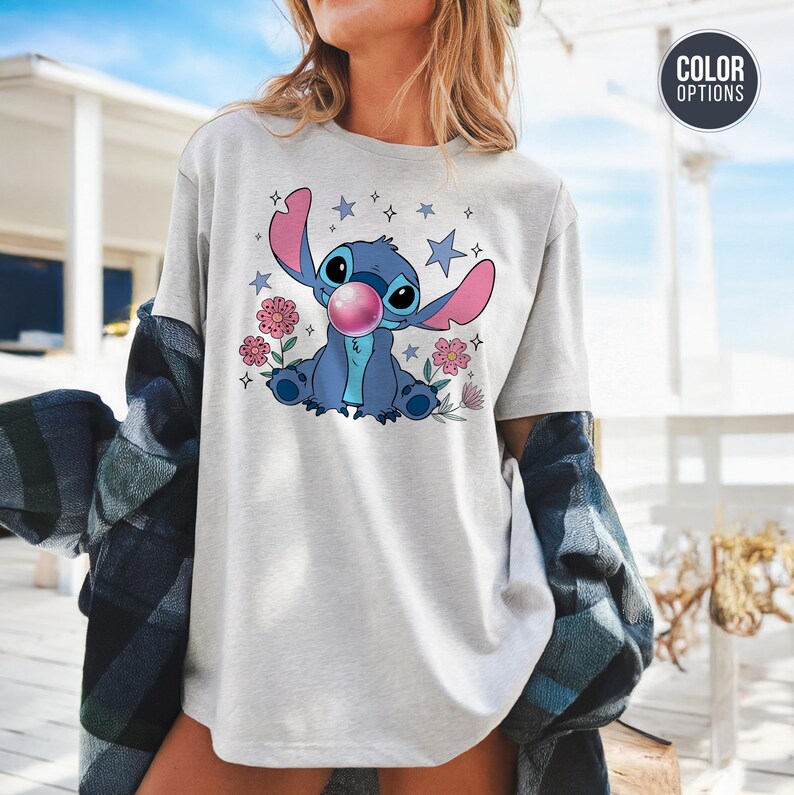 May include: A light gray t-shirt featuring a cartoon illustration of Stitch blowing a bubble. The blue Stitch is surrounded by pink flowers and stars. The shirt has a crew neck and short sleeves.