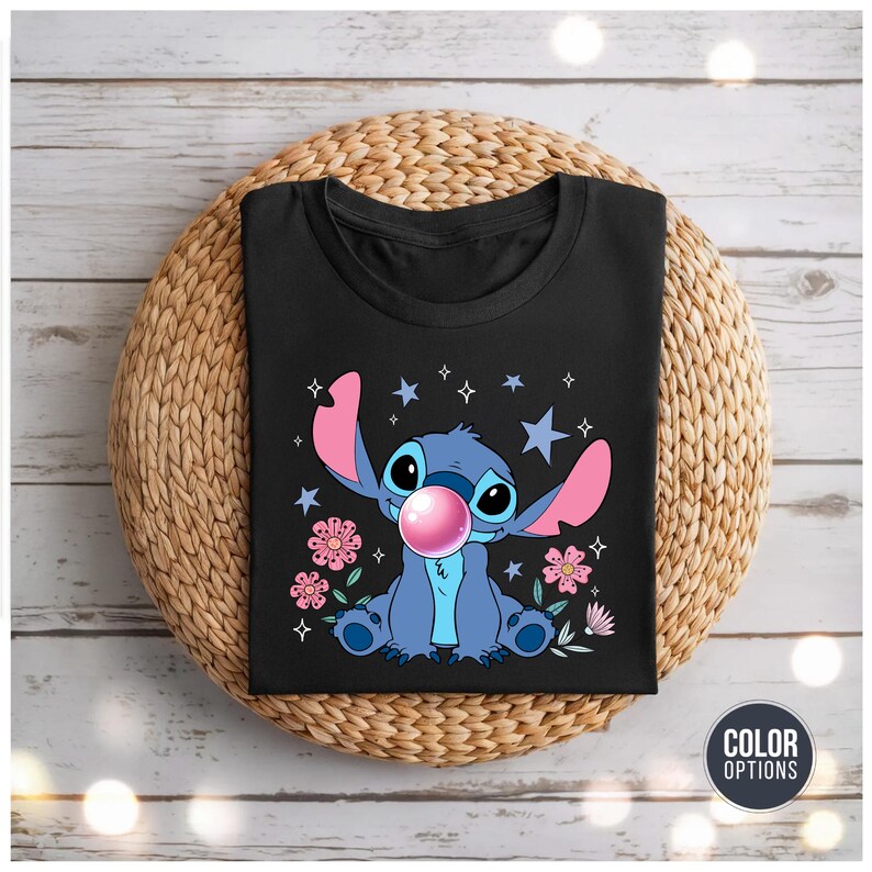May include: Black t-shirt featuring a cartoon character with blue fur, large ears, and a pink bubble gum bubble. The design includes pink flowers, stars, and the character's name. The shirt is displayed on a woven, circular mat.