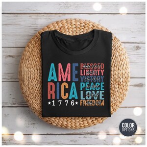 May include: Black t-shirt with the word "AMERICA" in colorful block letters. The shirt also features the text "BLESSED, LIBERTY, VICTORY, PEACE, LOVE, FREEDOM" and the year "1776". The shirt is displayed on a woven basket.