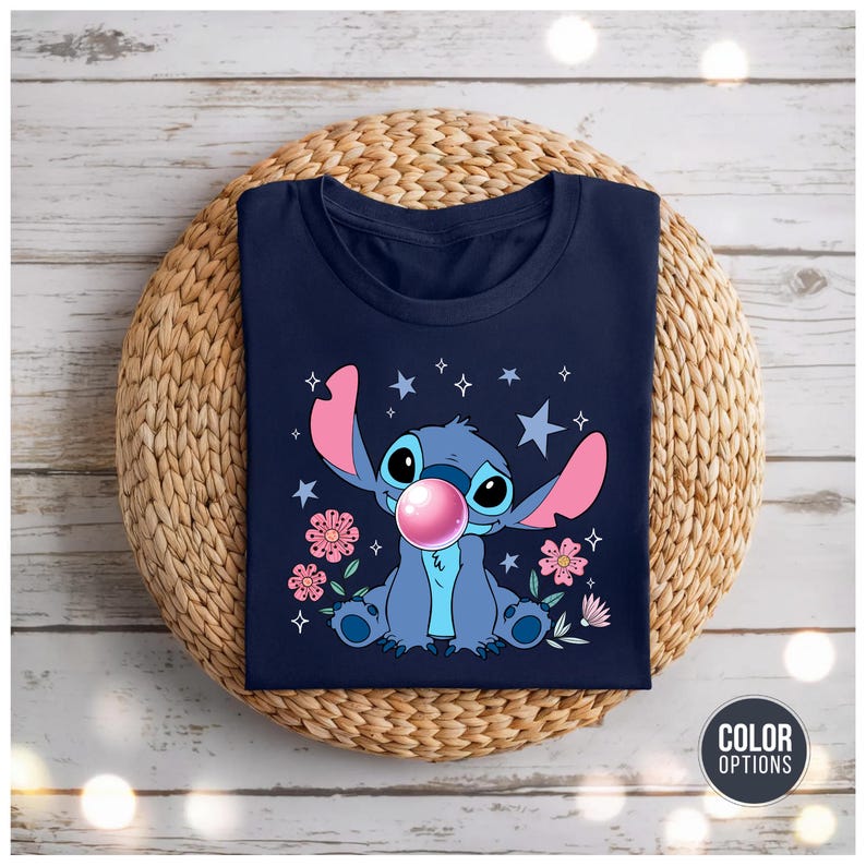 May include: Navy blue t-shirt featuring a cartoon illustration of Stitch blowing a bubble. The design includes pink flowers, stars, and a pink bubble. The shirt is displayed on a woven, circular mat.