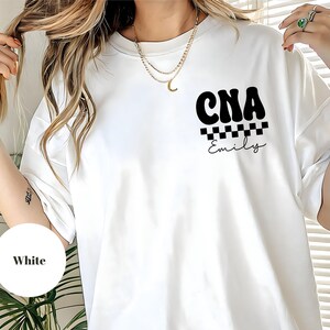 K&ouml;nnte beinhalten: Wei&szlig;es T-Shirt mit dem schwarzen Text "CNA" &uuml;ber einem schwarz-wei&szlig;en Schachbrettmuster und dem Namen "Emily". Das T-Shirt ist im l&auml;ssigen Stil gehalten.