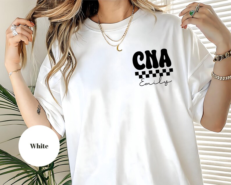 K&ouml;nnte beinhalten: Wei&szlig;es T-Shirt mit dem schwarzen Text "CNA" &uuml;ber einem schwarz-wei&szlig;en Schachbrettmuster und dem Namen "Emily". Das T-Shirt ist im l&auml;ssigen Stil gehalten.