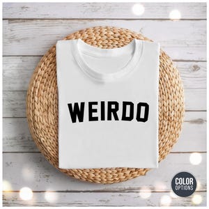 May include: A white t-shirt with the word "WEIRDO" printed in bold, black letters. The shirt is neatly folded and rests on a round, woven wicker placemat. The background features a light-colored wooden surface.