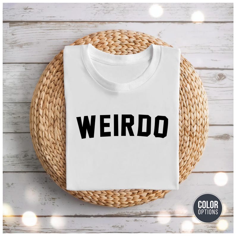 May include: A white t-shirt with the word "WEIRDO" printed in bold, black letters. The shirt is neatly folded and rests on a round, woven wicker placemat. The background features a light-colored wooden surface.