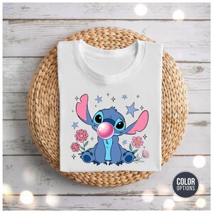 May include: White t-shirt featuring a blue cartoon character with large ears, blowing a pink bubble. The design includes pink flowers, blue stars, and sparkles. The shirt is displayed on a woven, circular surface.