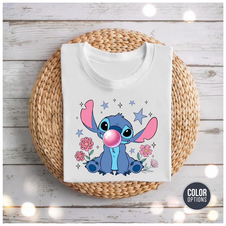 May include: White t-shirt featuring a blue cartoon character with large ears, blowing a pink bubble. The design includes pink flowers, blue stars, and sparkles. The shirt is displayed on a woven, circular surface.