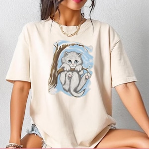 May include: Cream-colored t-shirt featuring a graphic of a gray kitten hanging from a tree branch. The kitten has blue eyes and pink paws, set against a light blue background with snowflakes. The shirt has short sleeves and a relaxed fit.