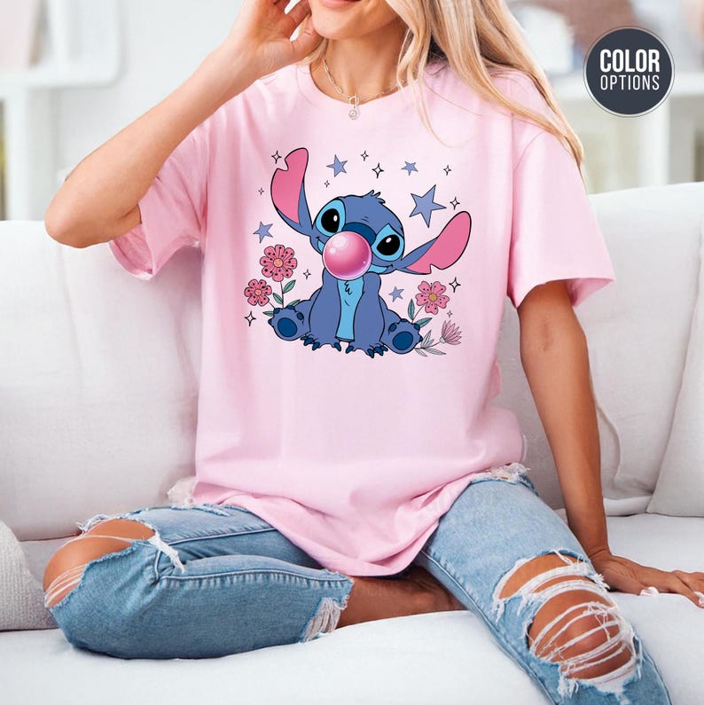 May include: Pink t-shirt featuring a blue cartoon character blowing a bubblegum bubble. The graphic includes stars and flowers. The shirt is a light pink color.
