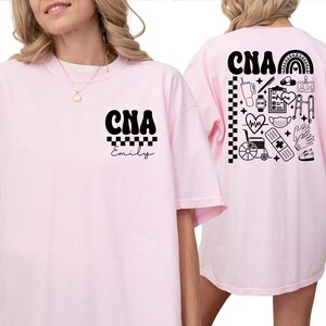 K&ouml;nnte beinhalten: Hellrosa T-Shirt mit den schwarzen Buchstaben "CNA" &uuml;ber dem Namen "Emily" in Schreibschrift. Auf der R&uuml;ckseite des Shirts befindet sich ein schwarz-wei&szlig;es Design mit medizinischen Symbolen, darunter ein Regenbogen, ein Herz und ein Stethoskop.