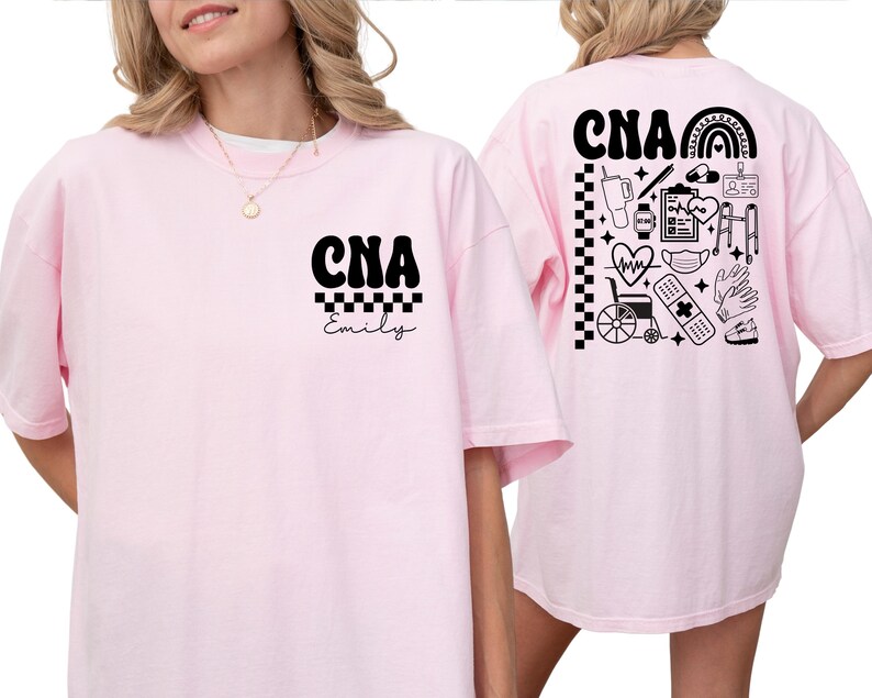 K&ouml;nnte beinhalten: Hellrosa T-Shirt mit den schwarzen Buchstaben "CNA" &uuml;ber dem Namen "Emily" in Schreibschrift. Auf der R&uuml;ckseite des Shirts befindet sich ein schwarz-wei&szlig;es Design mit medizinischen Symbolen, darunter ein Regenbogen, ein Herz und ein Stethoskop.