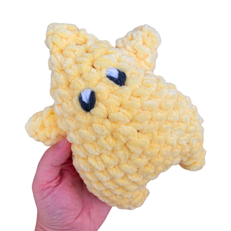 Puede incluir: Un peluche de ganchillo de color amarillo p&aacute;lido con forma de estrella. La estrella presenta una textura de lana gruesa y voluminosa, con dos ojos bordados, cada uno compuesto por un &oacute;valo azul marino oscuro con un peque&ntilde;o punto blanco.