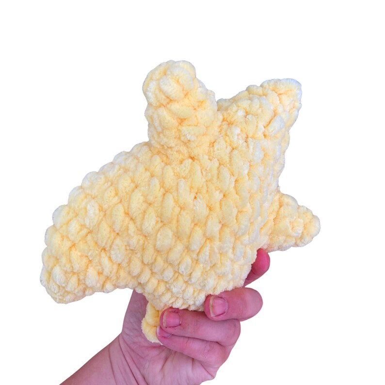 Puede incluir: Un juguete de peluche de color amarillo p&aacute;lido con forma de estrella de cinco puntas, elaborado con hilo grueso y texturizado con un aspecto esponjoso y rizado. La estrella presenta puntas redondeadas y una construcci&oacute;n densa y rellena.