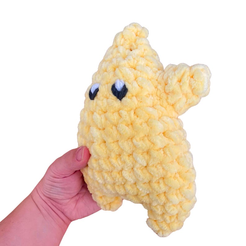 Puede incluir: Un peluche de ganchillo de color amarillo p&aacute;lido con forma de estrella de cinco puntas. El juguete est&aacute; elaborado con un hilo grueso y texturizado que muestra un patr&oacute;n de puntos en bucle, con dos ojos bordados en hilo negro y blanco.