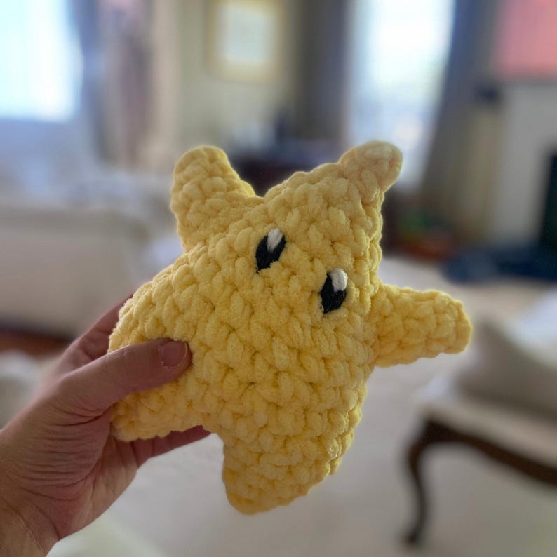 Puede incluir: Un peluche con forma de estrella amarilla hecho de hilo grueso. La estrella presenta una superficie texturizada de ganchillo con dos ojos bordados, cada uno compuesto por un &oacute;valo negro con un peque&ntilde;o acento blanco, d&aacute;ndole una expresi&oacute;n facial.