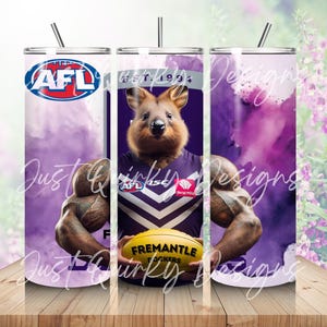 Puede incluir: Un vaso de acero inoxidable con un quokka que lleva una camiseta de los Fremantle Dockers, sosteniendo un balón de fútbol amarillo. El diseño incluye el logotipo de la AFL y efectos de humo púrpura. El vaso tiene una pajita negra.