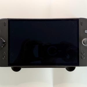 May include: A black handheld gaming console featuring a large rectangular screen, two analog joysticks, a directional pad, and four action buttons. The device has a smooth, rounded frame with integrated controls on both the left and right sides.