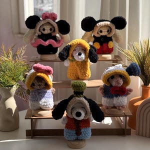 May include: A selection of handmade crochet dolls, each dressed in character-inspired outfits. The dolls feature designs like Mickey and Minnie Mouse, a yellow dog, and other cartoon characters. They are displayed on wooden stands.