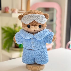 May include: A brown crocheted bear plush toy wearing a light blue pajama set with a collared shirt and matching shorts. The bear has black plastic eyes and a light blue sleep mask positioned over its forehead, all crafted from soft yarn.
