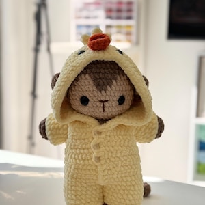 May include: A handmade crochet monkey doll wearing a yellow hooded jumpsuit with button closures. The monkey has brown accents on its ears and hands, and a small orange crown on its head. The doll is standing on a wooden base.