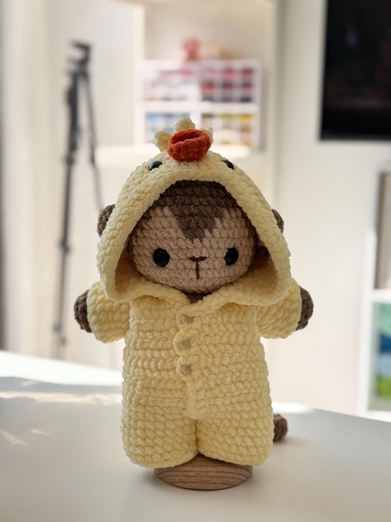 May include: A handmade crochet monkey doll wearing a yellow hooded jumpsuit with button closures. The monkey has brown accents on its ears and hands, and a small orange crown on its head. The doll is standing on a wooden base.