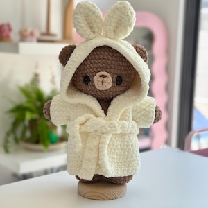 May include: A brown crocheted bear plush toy wearing a cream-colored hooded bathrobe with bunny ears. The bear has dark round eyes and a light tan muzzle. The plush is standing on a small, round, light-colored wooden base, showcasing a soft texture.