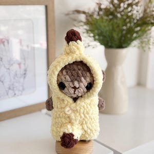 以下が含まれることがあります： 黄色いフード付きコートと茶色のトップスを着た、手作りのかぎ針編みのテディベア。クマは黒い目を持ち、木の台座の上に立っています。コートはシングルボタン留めです。クマは可愛くて柔らかいおもちゃです。