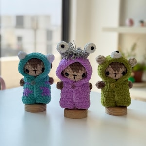 May include: Three small crocheted figures standing on wooden bases. The left figure wears a teal hooded cloak with purple spots. The center figure wears a purple hooded cloak with two white eyes and gray hair. The right figure wears a green hooded cloak.